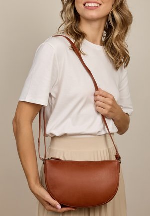 Woman wearing white shirt and beige skirt holds brown leather crossbody bag with adjustable strap and zipper closure.