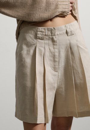 Beige hooggetailleerde geplooide korte broek gedragen met een cropped textuur gebreide trui, waarbij de buik en taillebanddetails zichtbaar zijn.