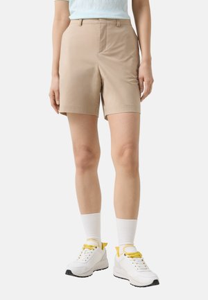 Person trägt beige knieumspielende Shorts, weiße Socken und weiße Turnschuhe mit gelben Schnürsenkeln und steht vor einem einfarbigen Hintergrund.