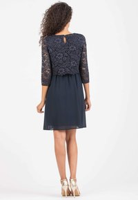 Marineblaues Kleid mit Spitzentop und 3/4-Ärmeln, mit einem Schlüsselrunde-Ausschnitt hinten und einem fließenden Chiffonrock. Goldene High Heels runden den Look ab.
