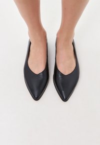 Des ballerines en cuir noir avec un design à bout pointu, une texture lisse et une silhouette en V, présentées de haut sur des pieds nus.