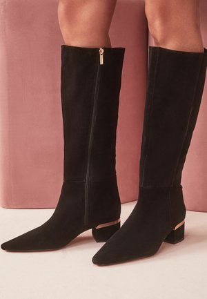 Botas negras hasta la rodilla con punteras puntiagudas, tacones bajos de bloque con detalles dorados, usadas por una persona de pie frente a un fondo rosa.