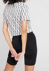 Eine Person trägt schwarze Shorts und ein weißes, gemustertes Shirt mit schwarzen geometrischen Formen und steht mit hinter dem Rücken verschränkten Händen.
