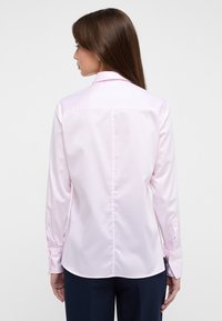 Camicia a maniche lunghe di un delicato rosa con colletto a punta, dettagli sul girocollo posteriore e polsini con bottoni, realizzata in tessuto liscio.