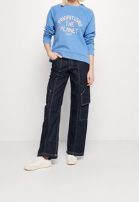 Blå sweatshirt med trycket "Prioritera planeten", kombinerad med mörka denim cargo-byxor med vit söm och flera fickor. Vita sneakers.