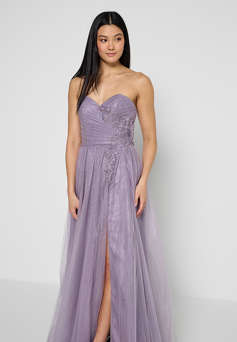Suknia w kolorze lawendy bez ramiączek, z dopasowanym gorsetem, ozdobnymi kwiatowymi detalami oraz zwiewną spódnicą z rozcięciem do uda i lśniącą teksturą.