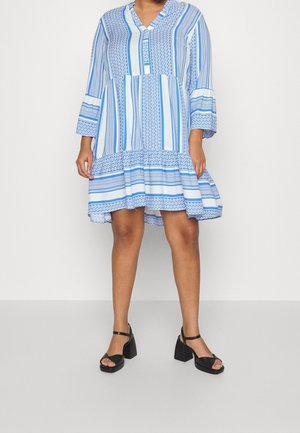 Frau trägt ein blau-weiß gemustertes knielanges Kleid mit langen Ärmeln und schwarzen offenen Sandalen mit Blockabsatz und steht vor einem schlichten Hintergrund.