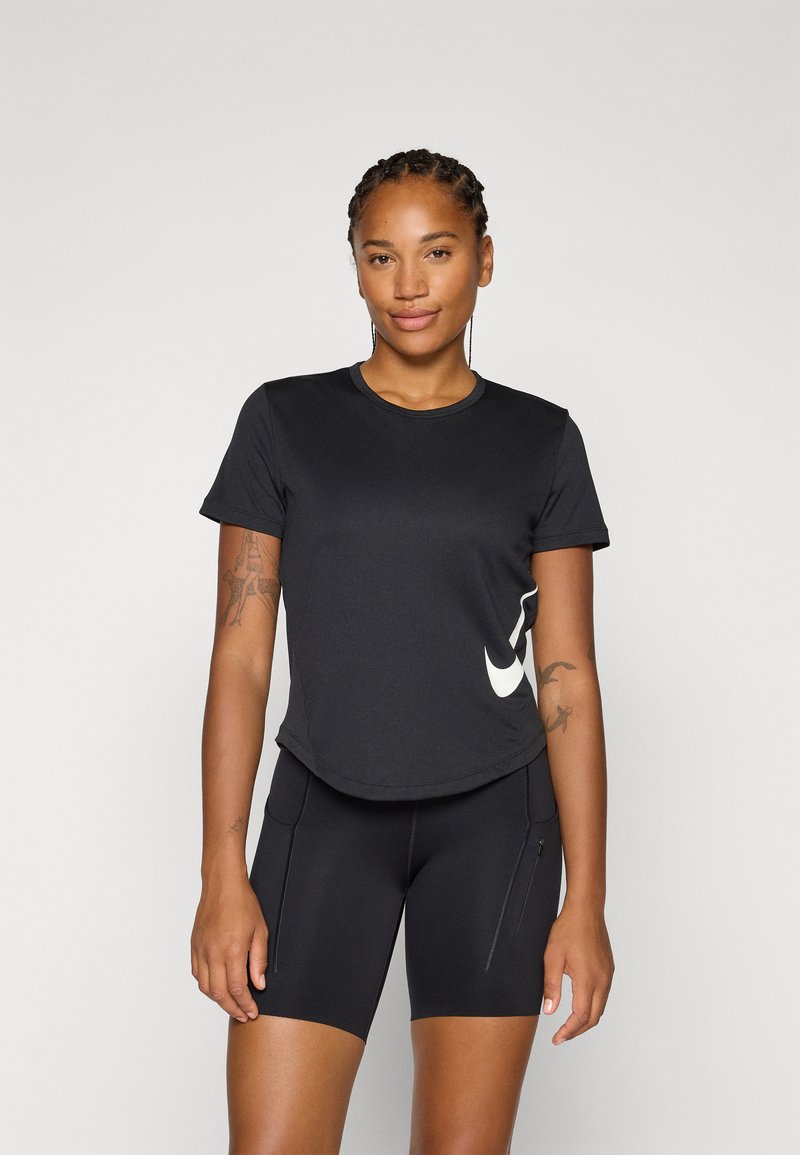 Femme souriante avec des cheveux tressés portant un haut de sport noir et un short assorti, affichant un logo sur son T-shirt, sur un fond clair.