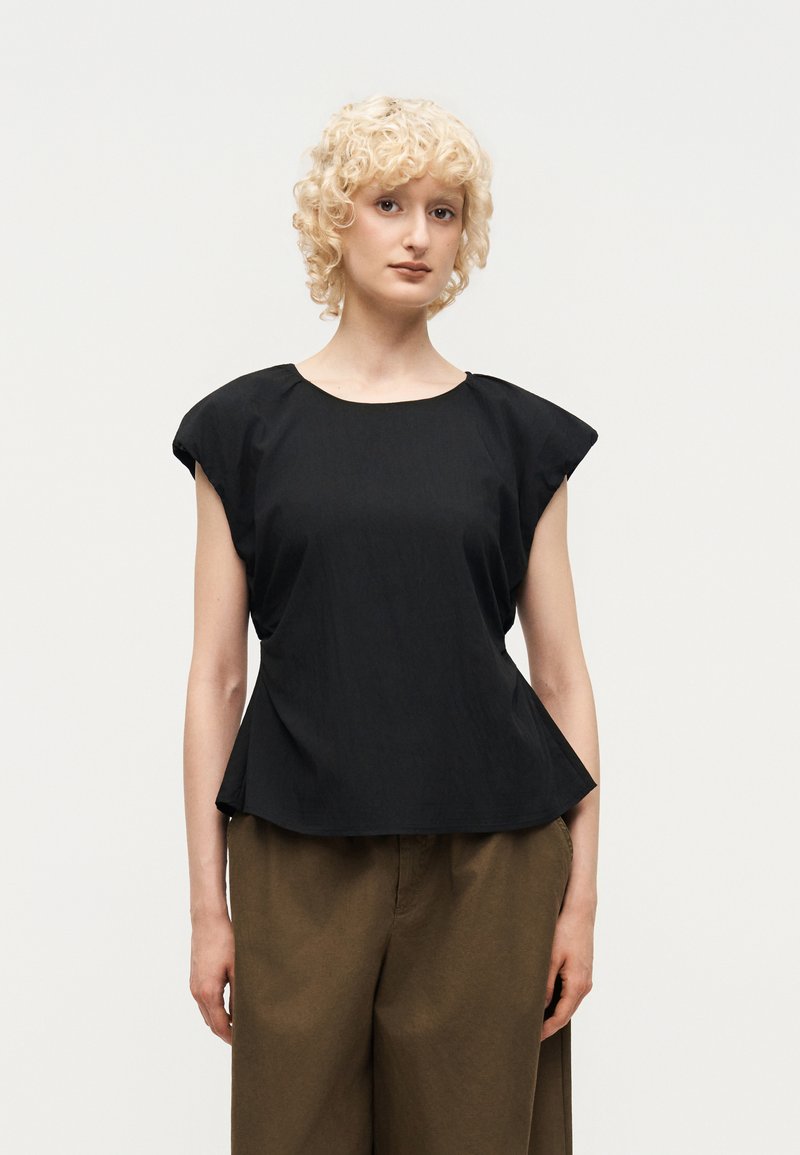 Jeune personne aux cheveux blonds courts et bouclés portant un haut noir sans manches avec des épaules rembourrées et un pantalon marron ample, debout sur fond blanc.
