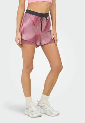 Nő rózsaszín-fehér csíkos futónadrágot visel fekete derékpánttal, rózsaszín belső réteggel, fehér zoknival és fehér sportcipővel.