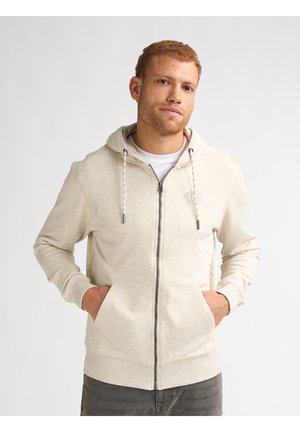 Man met een licht beige hoodie met rits en handen in de voorzakken, staand tegen een effen witte achtergrond.