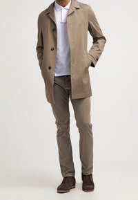 Beige lichtgewicht jas met drie knopen, gelaagd over een witte polo. Gecombineerd met lichtbruine broek en donkerbruine schoenen. Glad textuur.