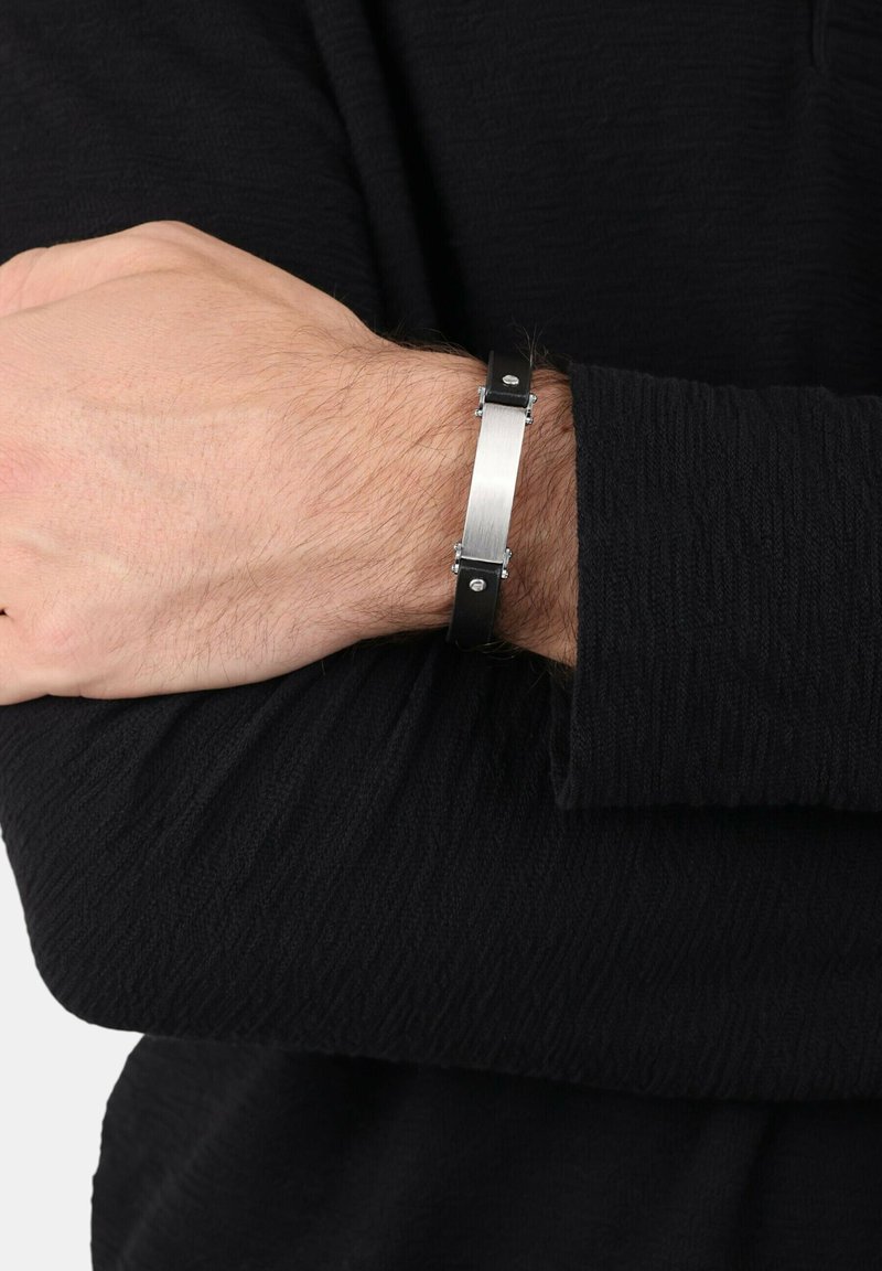 Muñeca masculina cruzada sobre el torso, llevando una pulsera de cuero negra con una placa metálica rectangular plateada y remaches plateados.