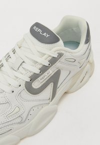 Zapatilla blanca con acentos grises, parte superior texturizada y suela acolchada; cuenta con un diseño de cordones y un logo en el talón y el costado.