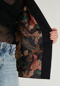 Černý blazer s vzorovaným interiérem, který obsahuje paisley v červené, zelené a zlaté na tmavém pozadí. Džíny z denimu jsou viditelné v pase.