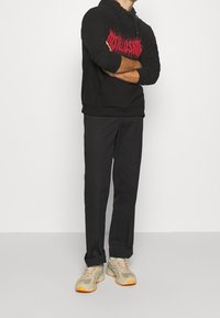 Černá mikina s kapucí a červeným grafickým textem, spárovaná s černými kalhotami a béžovými teniskami s oranžovými akcenty. Hladká tkanina a uvolněný střih.