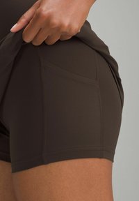 Mano levantando un pantalón corto atlético de color marrón oscuro, con el bolsillo en la parte superior del muslo, mostrando un primer plano de los detalles de la tela y las costuras.