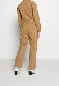 Jumpsuit de algodón en tono marrón claro con mangas largas, bolsillos traseros y un ajuste holgado. Presenta un cuello alto y puños con botones, combinados con botas blancas de tacón.