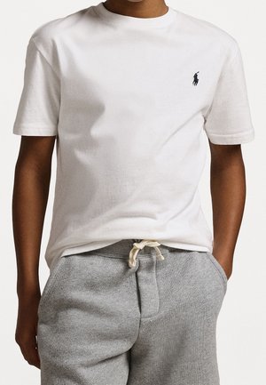 Jongen draagt een simpel wit T-shirt met korte mouwen en een klein zwart logo op de borst en grijze joggingbroek met trekkoord, handen in de zakken.
