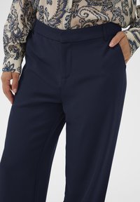 Osoba v tmavomodrých upravených nohaviciach s rukami v vreckách a dlhým rukávom tričkom s béžovým paisley vzorom zapnutým dovnútra.