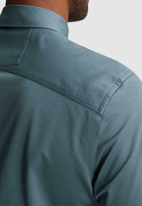 Homme portant une chemise bleu-vert avec « Cast Iron » brodé sur le haut du dos près de la couture de l’épaule.