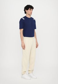 T-shirt bleu marine à manches courtes avec des accents rayés blancs sur les épaules, associé à un pantalon de survêtement couleur crème et des baskets blanches.