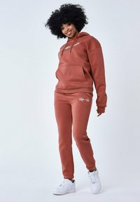 Sudadera color óxido y pantalones de chándal a juego en material suave, que presentan un bolsillo frontal y un detalle de logo. Zapatillas blancas completan el look.