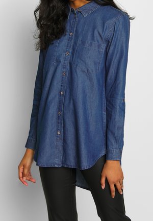 Femme portant une chemise en jean bleue à manches longues avec des poches poitrine et un pantalon noir, debout devant un fond uni.