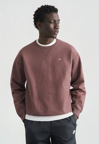 Μπορντό φούτερ με στρογγυλή λαιμόκοψη και λευκό λογότυπο Nike. Διαθέτει υφή επιφάνειας και άνετη εφαρμογή, φορεμένο πάνω από ένα λευκό πουκάμισο.