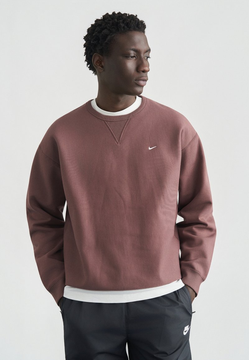 Μπορντό φούτερ με στρογγυλή λαιμόκοψη και λευκό λογότυπο Nike. Διαθέτει υφή επιφάνειας και άνετη εφαρμογή, φορεμένο πάνω από ένα λευκό πουκάμισο.