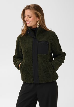 Femme souriante aux longs cheveux porte une veste polaire vert foncé avec une poche poitrine noire et un pantalon noir, les mains dans les poches de la veste.