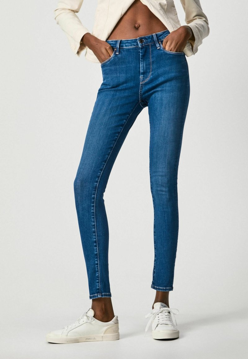 Skinny jeans in dark blue denim, featuring a high waist, two front pockets, and a zipper fly. Accompanied by white sneakers.