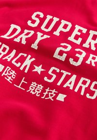 Červené bavlněné tričko s velkým, popraskaným bílým nápisem "SUPER DRY 23 TRACK STARS" vpředu, s dalšími znaky níže.