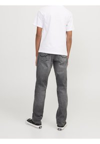 Persona con vaqueros grises y zapatillas negras y blancas, de pie de espaldas contra un fondo claro y liso.