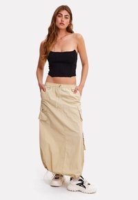 Femme portant un haut court noir sans bretelles, une jupe longue cargo beige avec ourlet à cordon de serrage, et des baskets blanches massives, debout les mains dans les poches.