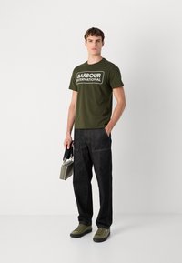 Khaki zelené tričko s potiskem "BARBOUR INTERNATIONAL" v bílé; tmavé džínové kalhoty; olivově zelené tenisky; nese šedou kabelku.