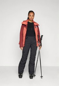 Skidräkt med en röd och orange isolerad jacka med huva, svart termotröja, svarta byxor och skidstavar. Texturerade tyger och figursydd design.