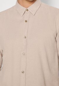 Jasnobeżowa koszula na guziki wykonana z teksturowanej tkaniny, z klasycznym kołnierzykiem i pięcioma guzikami z przodu.