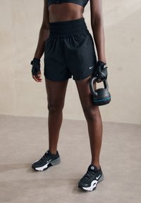 Pantalones cortos deportivos negros con una cinturilla elástica alta, combinados con zapatillas negras y blancas. Se sostiene una kettlebell negra en una mano.