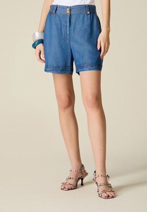 Kvinde iført blå denimshorts, hvid top, hælsandaler med slangeprint og chunky armbånd på venstre håndled, stående mod en cremefarvet baggrund.