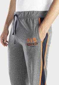 Graue Sweatpants mit einer entspannten Passform, die marineblaue Seitenstreifen und orangefarbene Schriftzüge aufweist. Sie sind mit einem Bund mit verstellbarem Kordelzug und Taschen ausgestattet.