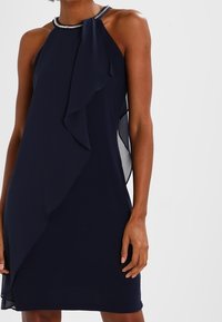 Robe halter bleu marine avec un vol froncé, des panneaux latéraux transparents et un détail de col perlé, fabriquée à partir d'un tissu lisse avec une silhouette ajustée.