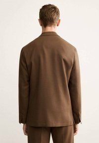 Blazer marrone con rever a punta, spalle strutturate e due bottoni su ciascun polsino; il tessuto appare liscio con una vestibilità sartoriale.