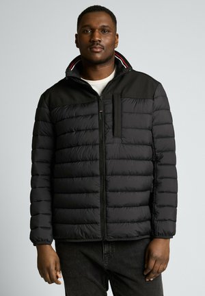 Mann trägt eine schwarze gesteppte Jacke mit Reißverschluss vorne und hohem Kragen, steht vor einem schlichten hellgrauen Hintergrund.