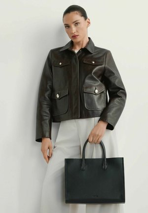 Kobieta w czarnej skórzanej kurtce i białych spodniach trzyma dużą czarną skórzaną torebkę z dwoma rączkami na jednolitym tle.