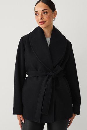 Vrouw met sproeten draagt een zwarte wollen jas met ceintuur en brede revers, gecombineerd met een zwarte broek, staand tegen een effen lichte achtergrond.