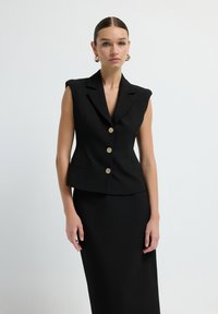Blazer sans manches noir à coupe ajustée, avec des boutons dorés et des revers nets. Tissu lisse au design minimaliste, assorti à une jupe coordonnée.