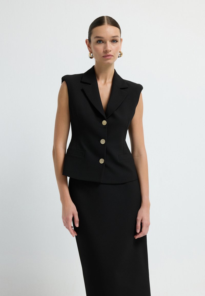 Blazer sans manches noir à coupe ajustée, avec des boutons dorés et des revers nets. Tissu lisse au design minimaliste, assorti à une jupe coordonnée.