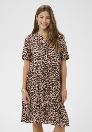 Femme souriante aux longs cheveux bruns portant une robe léopard à manches courtes, longueur genou, avec une ceinture à la taille et des bijoux en or, sur un fond clair.