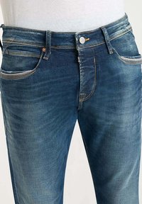 Vaqueros de mezclilla azul con un diseño clásico de cinco bolsillos, ligeras desgastaduras, cremallera y costuras en contraste.